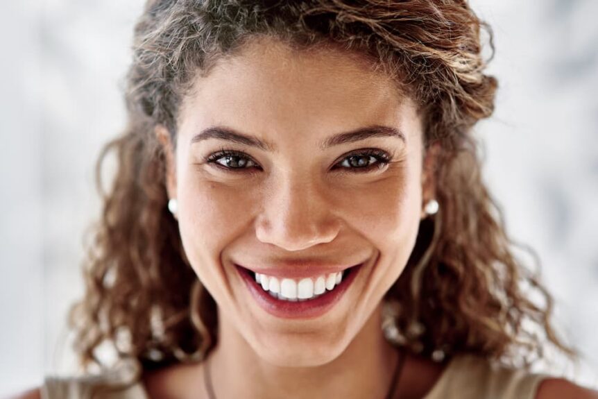 This image features a close-up portrait of a smiling young woman with curly hair. She appears to be happy and confident, showcasing a bright, healthy smile with white teeth after getting her teeth professionally whitened. Her open and friendly expression suggests a positive and cheerful demeanor.