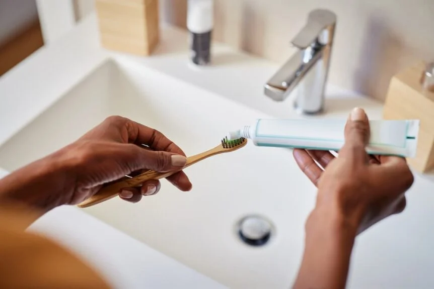 A person is preparing to brush their teeth, squeezing toothpaste onto an eco-friendly bamboo toothbrush in a modern bathroom.