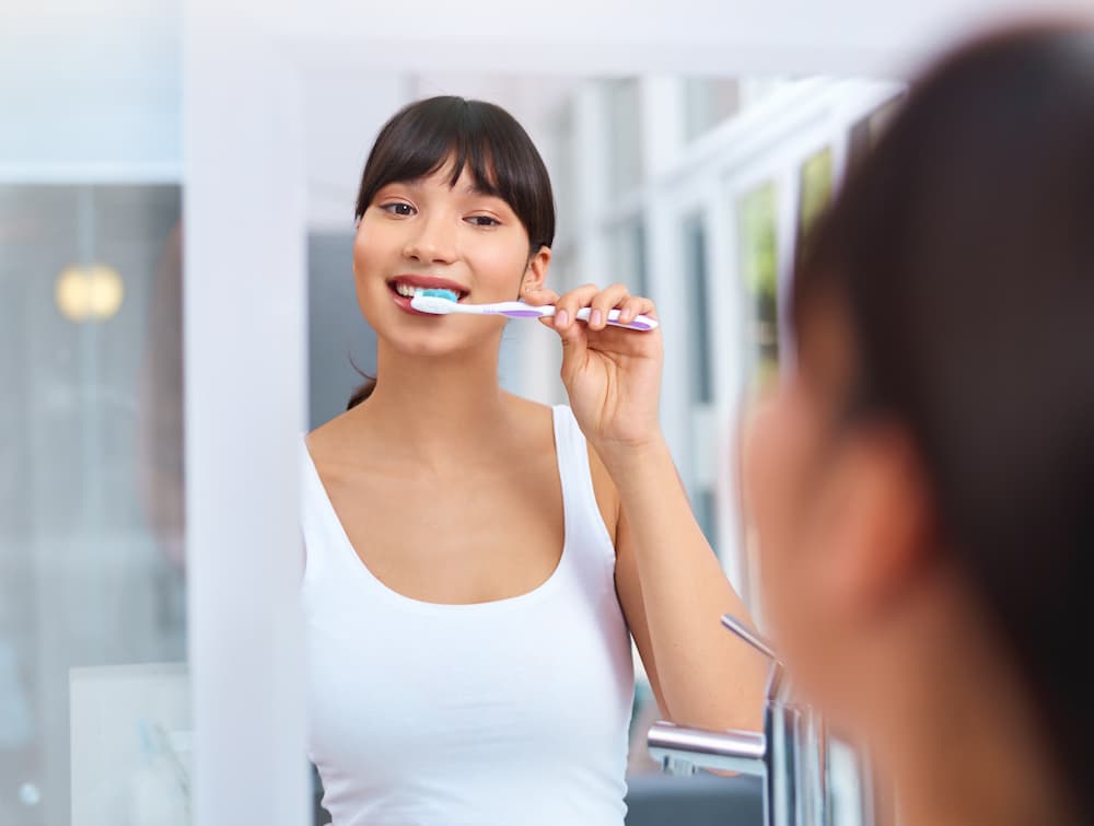 Young woman brushing her teeth with a toothbrush, applying toothpaste, in front of a mirror, reflecting a healthy dental routine.